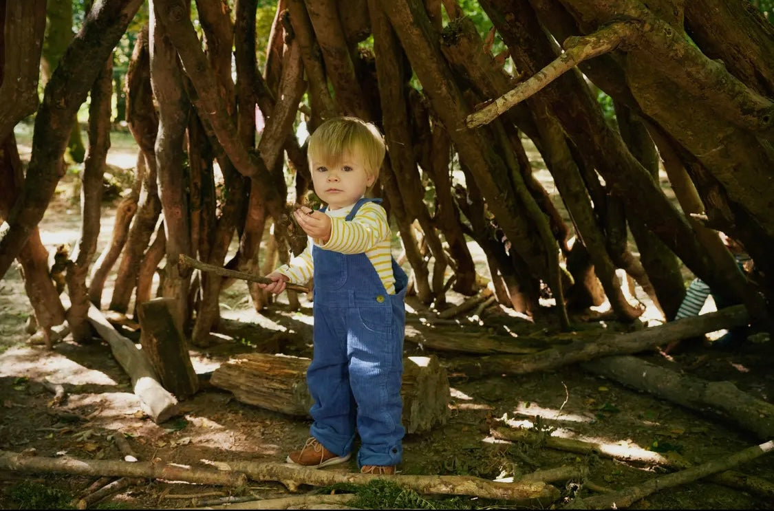 young child standing in tree branch hut holding stick wearing kids dotty dungaree breton yellow ochre white stripes bold white collar cotton jersey mix shoulder poppers snaps