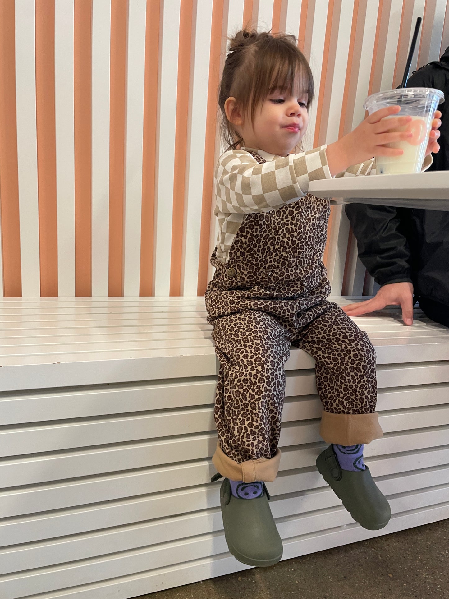 young child sitting on bench with drink wearing kids 100 % cotton dotty dungaree leopard brown tan animal print overalls adjustable straps rolled cuffs button cuffs 