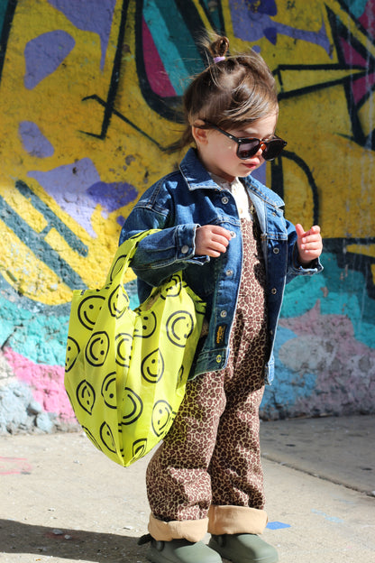 small child wearing blue denim jacket leopard overalls holding bright yellow smiley face reusable bag mini small baby baggu 100% recycled nylon 